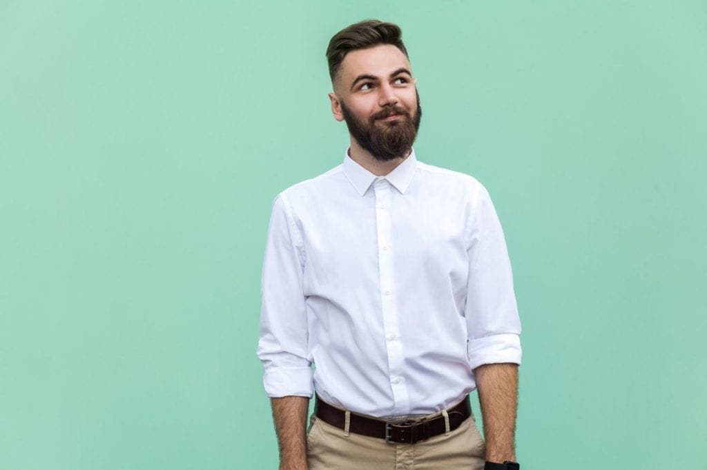 Taper comb over haircut: Brunhårig man med en borstad frisyr, bär en vit skjorta i beige byxor och står framför en mintgrön bakgrund